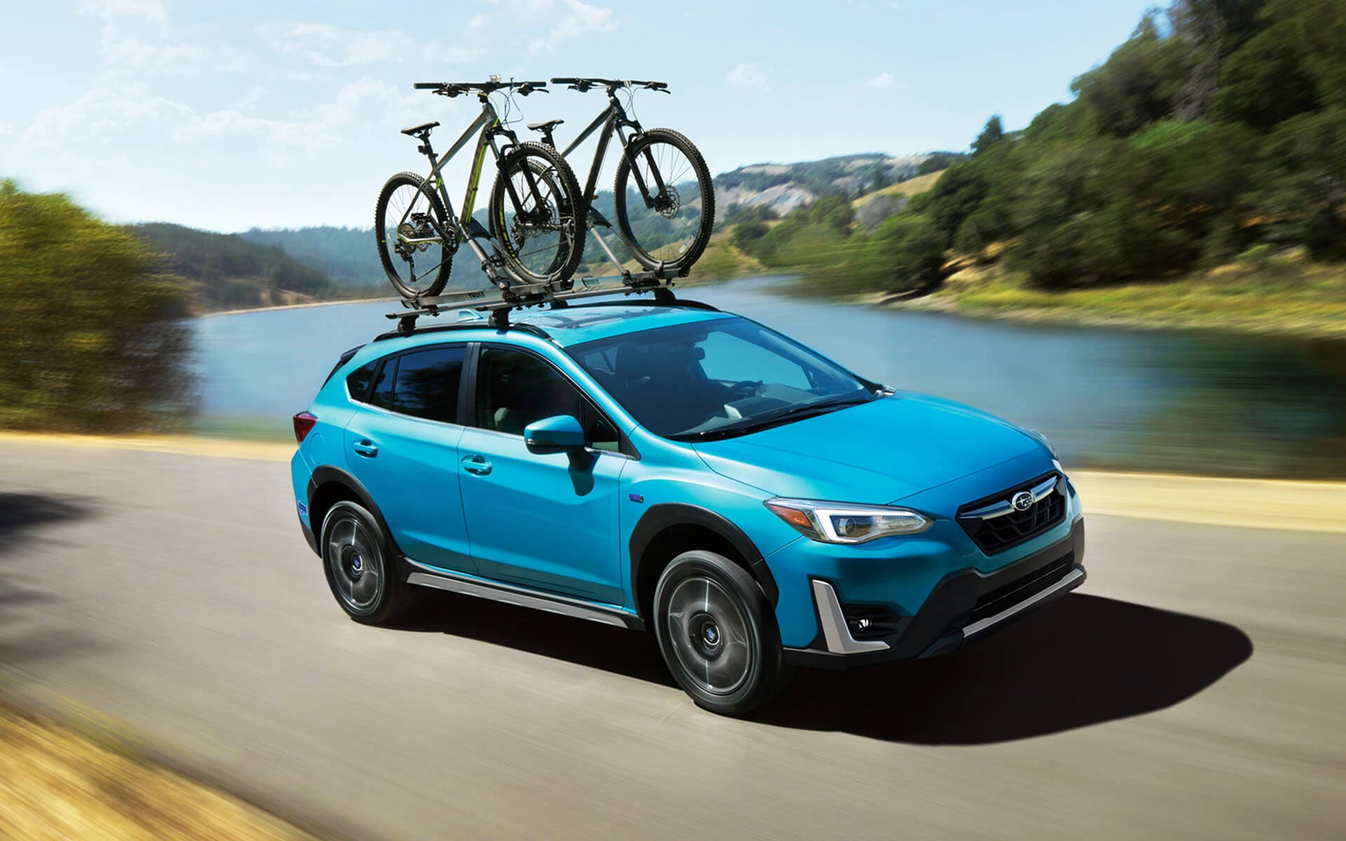 A blue Crosstrek Hybrid with two bicycles on its roof rack driving beside a river | Paul Moak Subaru in Jackson MS